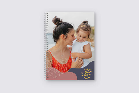 Une femme en haut rouge tient un enfant, debout à un bel endroit. La couverture du carnet montre des motifs floraux et des couleurs harmonieuses.